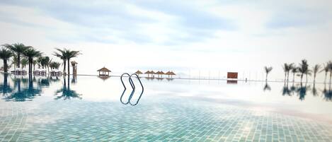 An infinity pool with clear blue tiles, palm trees, and beach huts in the background.