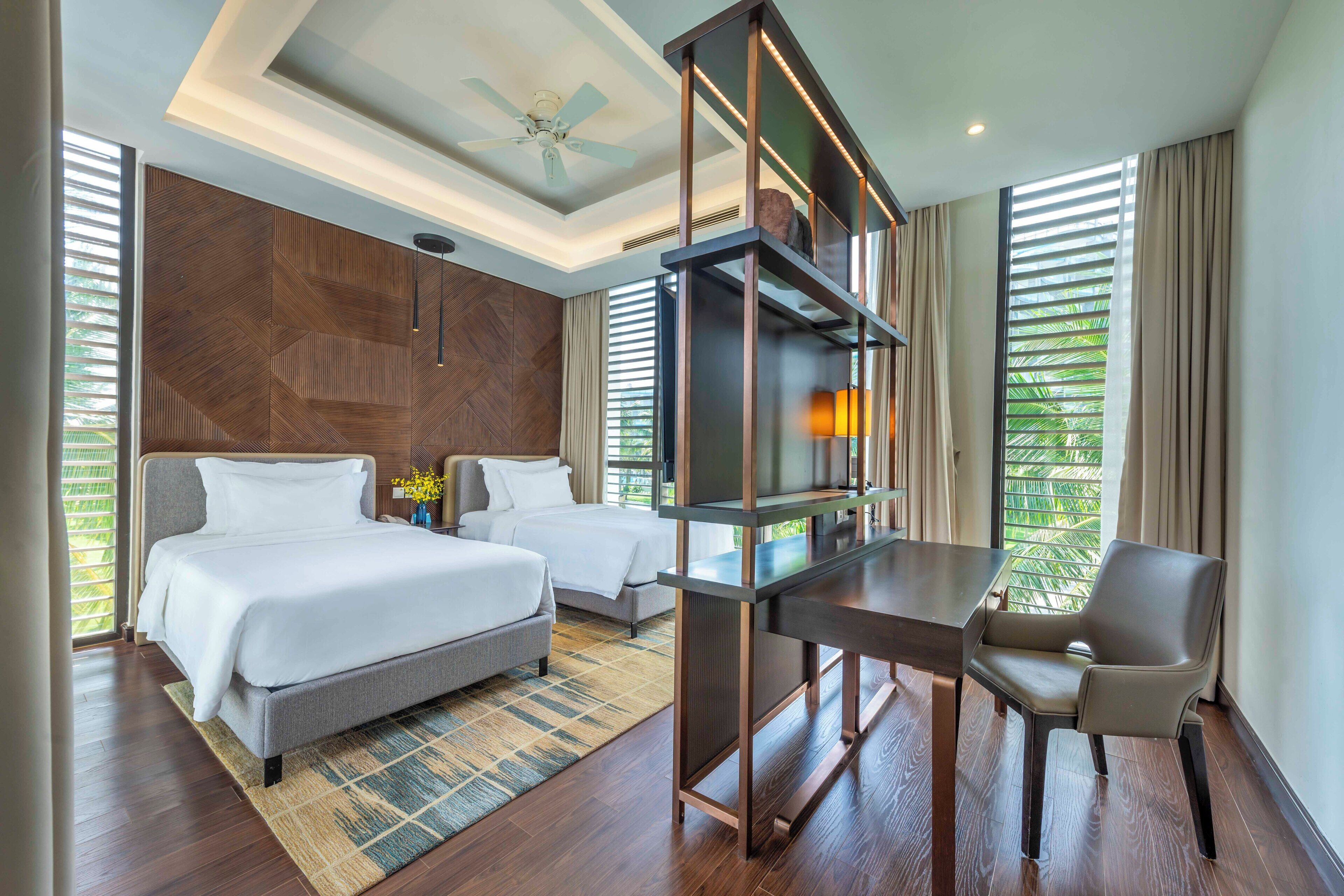 A hotel room with two beds, a desk, a chair, a ceiling fan, and a large window with blinds.