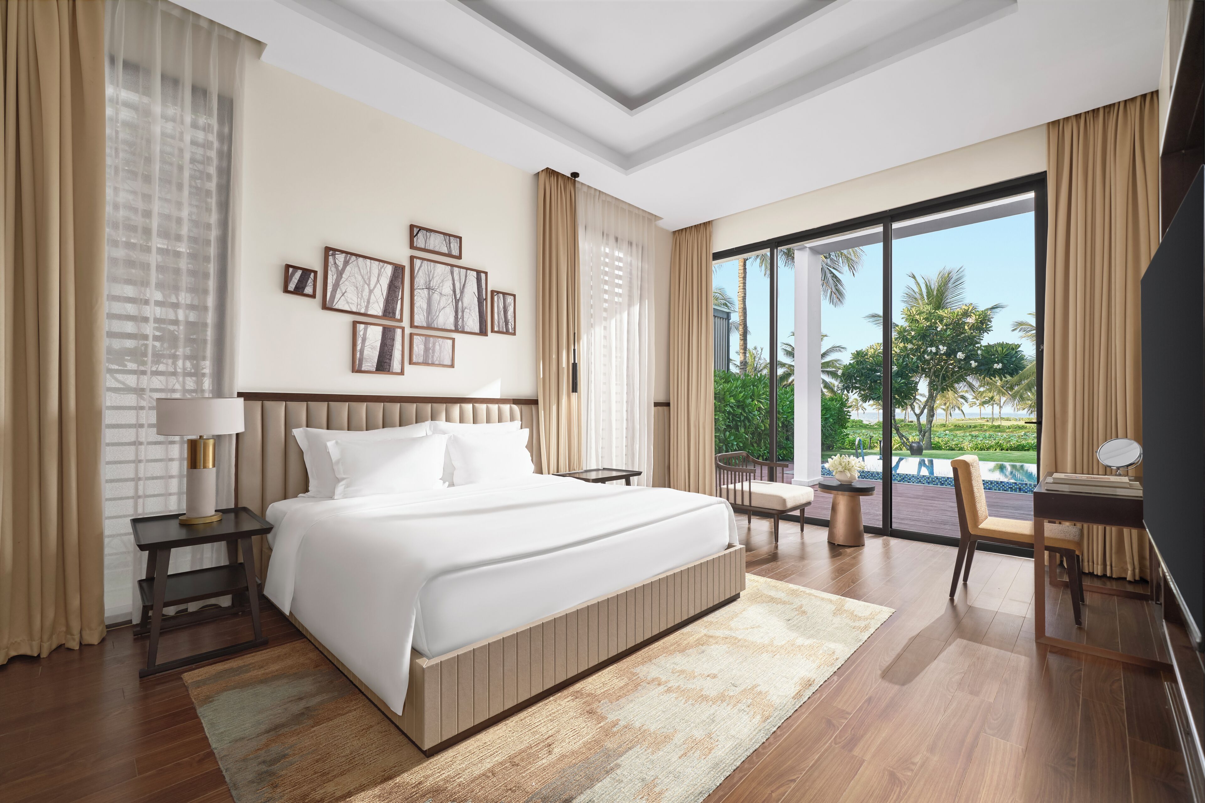 Ein Hotelzimmer mit einem großen Bett, einem Schreibtisch mit Stuhl, Blick auf Pool und Grünflächen sowie gerahmten Bildern an der Wand.
