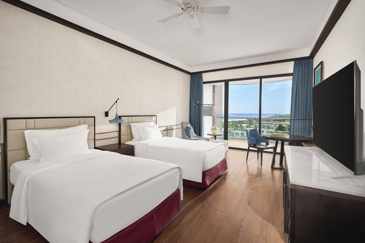 Een hotelkamer met twee bedden, een bureau, een stoel, een televisie en uitzicht op de oceaan.