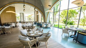 Un vaste espace de restauration intérieur, doté de tables rondes et de chaises en osier, de grandes fenêtres offrant une vue sur la verdure et d’un éclairage moderne suspendu.