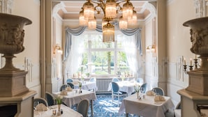 Une salle à manger élégante, avec des tables dressées pour le service, un grand lustre et une vue par la fenêtre.