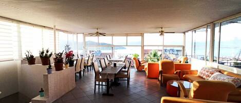 A spacious indoor area with a dining table, chairs, and orange sofas. There are potted plants on a ledge and a view of the sea through large windows.