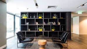A modern lobby with a large bookshelf, two leather chairs, a round wooden table, and a large window. - The William Suites (Melbourne)
