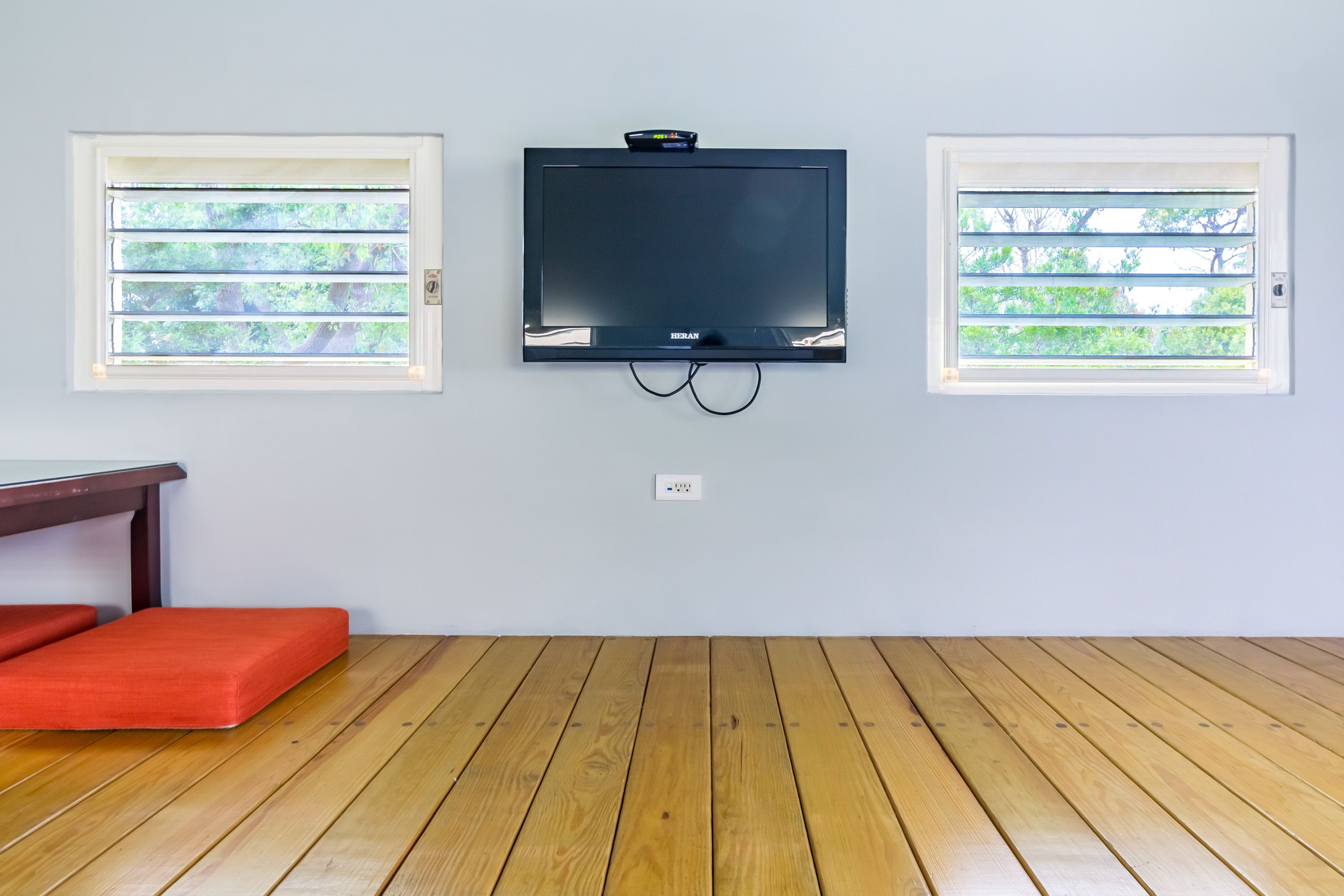 A room with a flat-screen TV mounted between two windows with blinds, a wooden floor, and a red bench.