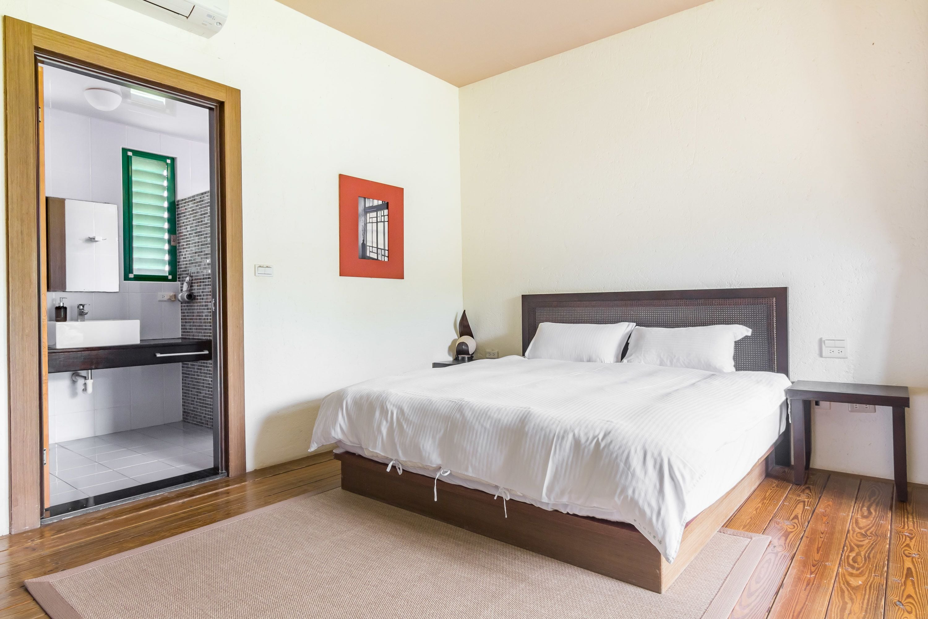 A bedroom with a bed, a bedside table, a nightstand, a mirror, a window with blinds, and a bathroom visible through an open door.