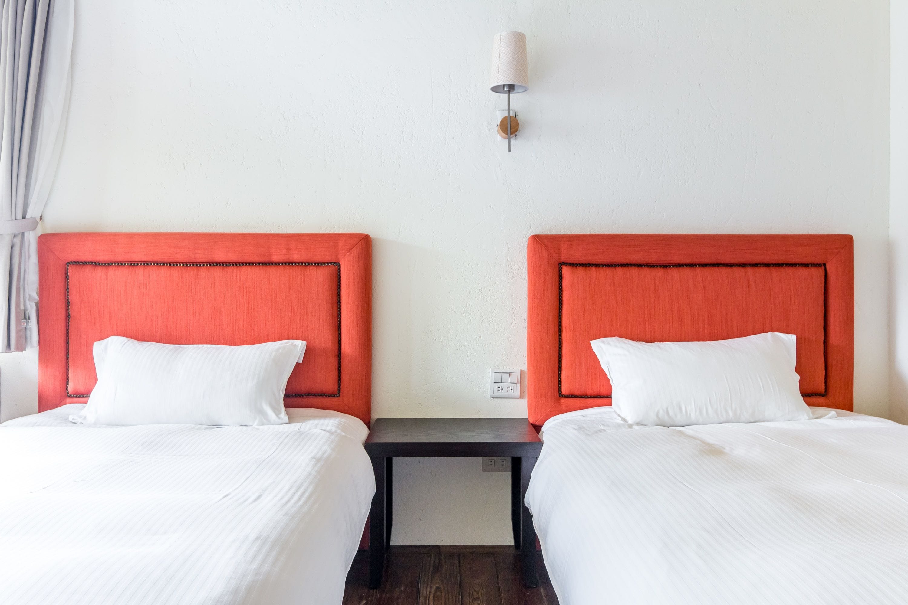 Habitación de hotel con dos camas, cada una con cabecero rojo y ropa de cama blanca. Hay una pequeña mesa entre las camas y una lámpara fijada a la pared encima.