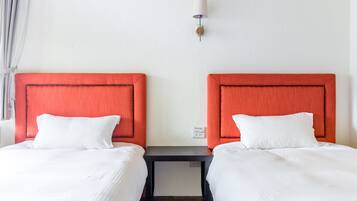 Habitación de hotel con dos camas, cada una con cabecero rojo y ropa de cama blanca. Hay una pequeña mesa entre las camas y una lámpara fijada a la pared encima.