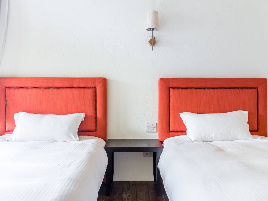 Habitación de hotel con dos camas, cada una con cabecero rojo y ropa de cama blanca. Hay una pequeña mesa entre las camas y una lámpara fijada a la pared encima.