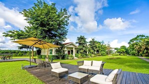 Une terrasse en bois, meublée de meubles en osier et agrémentée d’un parasol jaune, entourée d’une végétation luxuriante et avec une maison en arrière-plan.