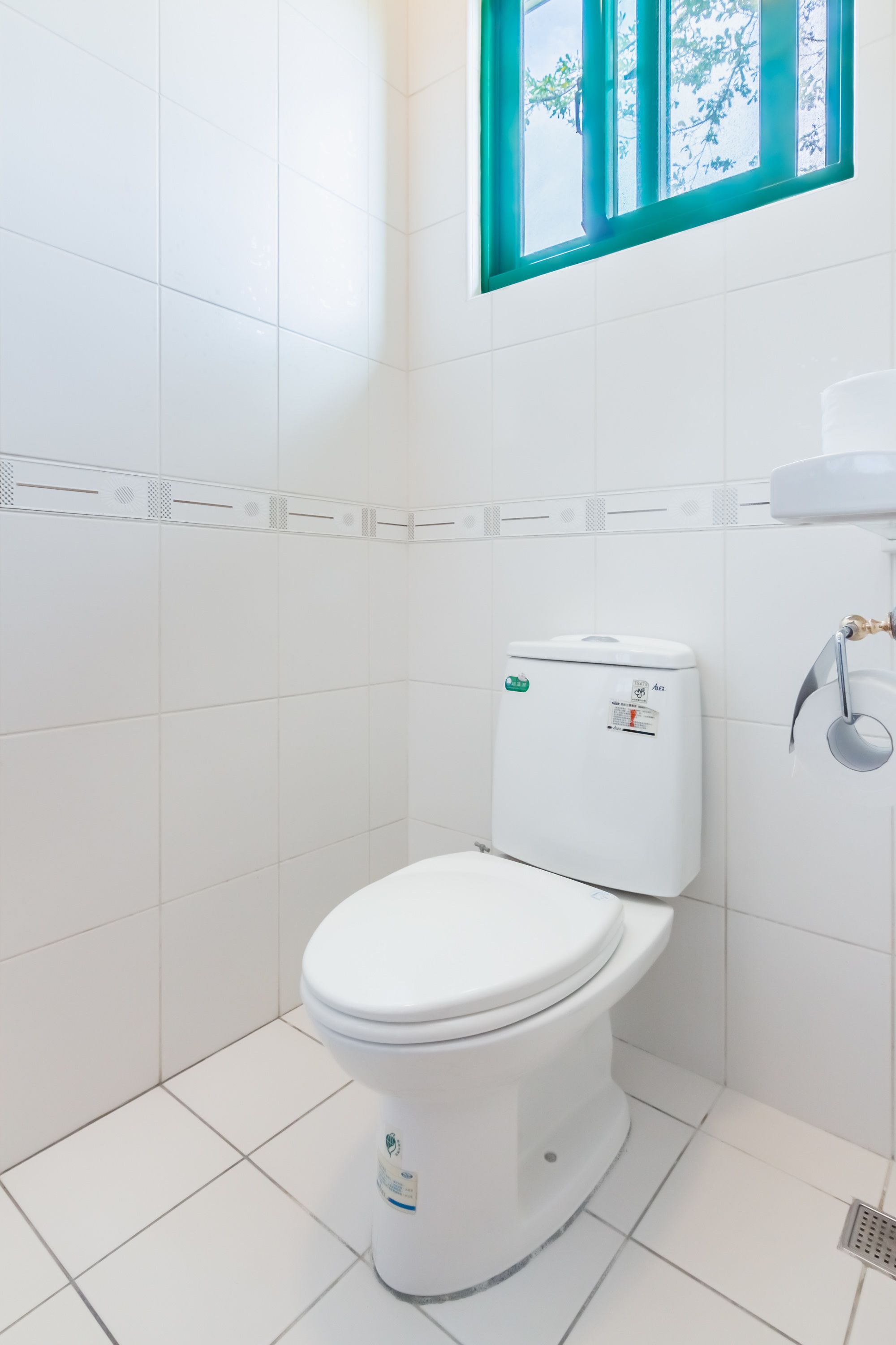 A white toilet with a tank, a window with a green frame, and a white tiled wall.