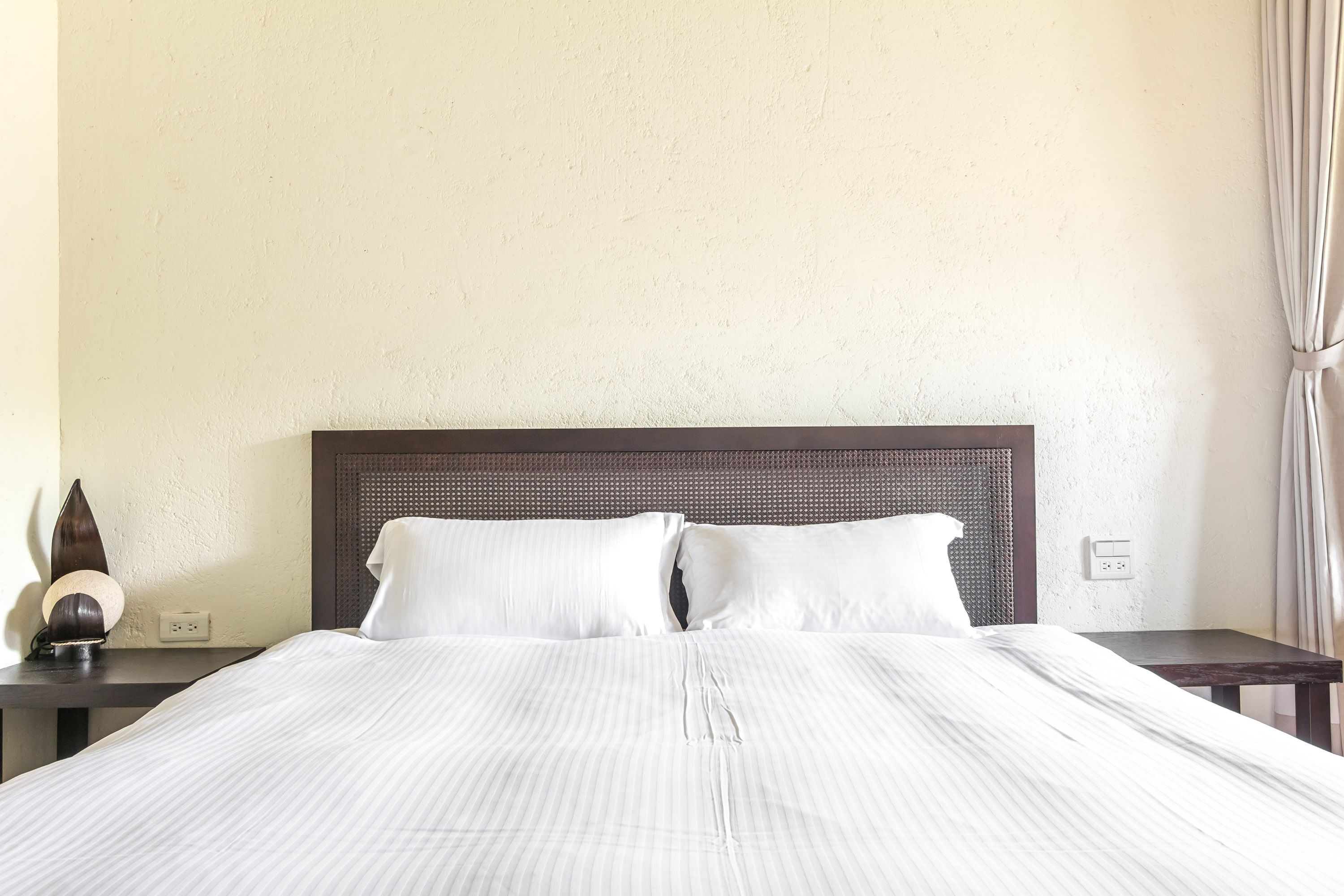 Una cama bien hecha con sábanas blancas, cabecera oscura y mesita de noche con lámpara y un objeto decorativo.