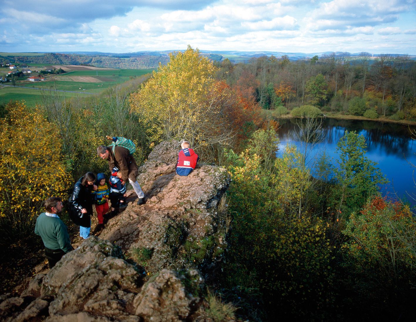 A group of people hiking on a rocky path with a lake and autumn foliage in the background.