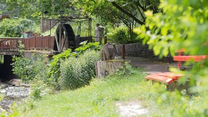 Un puente de madera rústico cruzando un arroyo con una rueda hidráulica y una estatua de una figura al fondo.