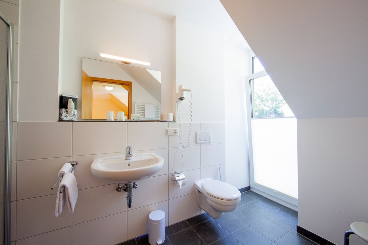 Een moderne badkamer met een wit wastafel, spiegel en toilet. Een handdoekrek met handdoeken, een prullenbak en een haardroger zijn aan de muur bevestigd.