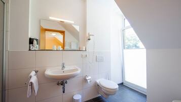 Een moderne badkamer met een wit wastafel, spiegel en toilet. Een handdoekrek met handdoeken, een prullenbak en een haardroger zijn aan de muur bevestigd.
