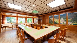 Un salón de conferencias con una larga mesa y sillas, ventanas amplias con vista a un área boscosa y un techo de madera con iluminación empotrada.