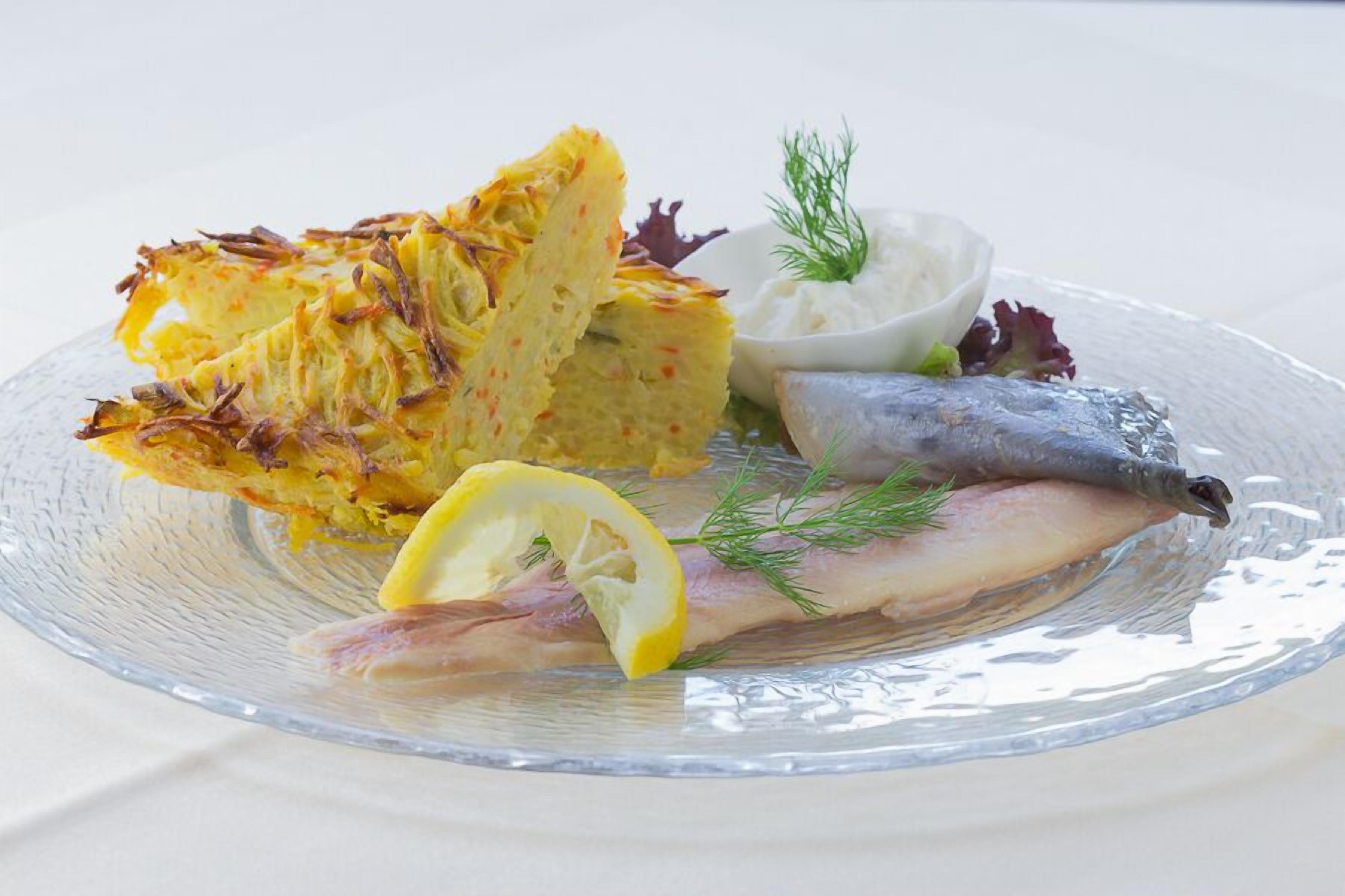 Una fuente de vidrio transparente con una porción de ensalada de patatas, un trozo de pescado, un gajo de limón y un pequeño recipiente con salsa blanca, decorada con eneldo.