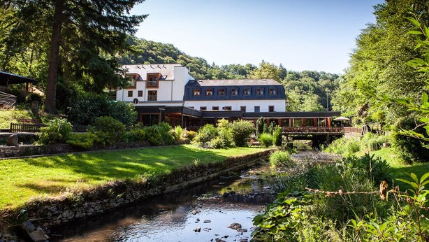 Un hotel a orillas del río con un canal revestido de piedra, abundante vegetación y un puente que conecta los edificios.