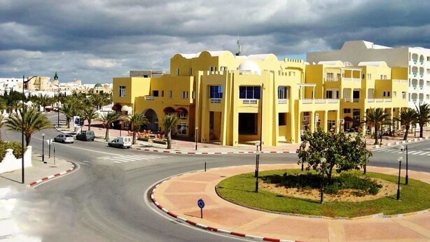 A yellow building with a central entrance and multiple windows, surrounded by palm trees and a roundabout. - Residence Aladin (Hammamet)