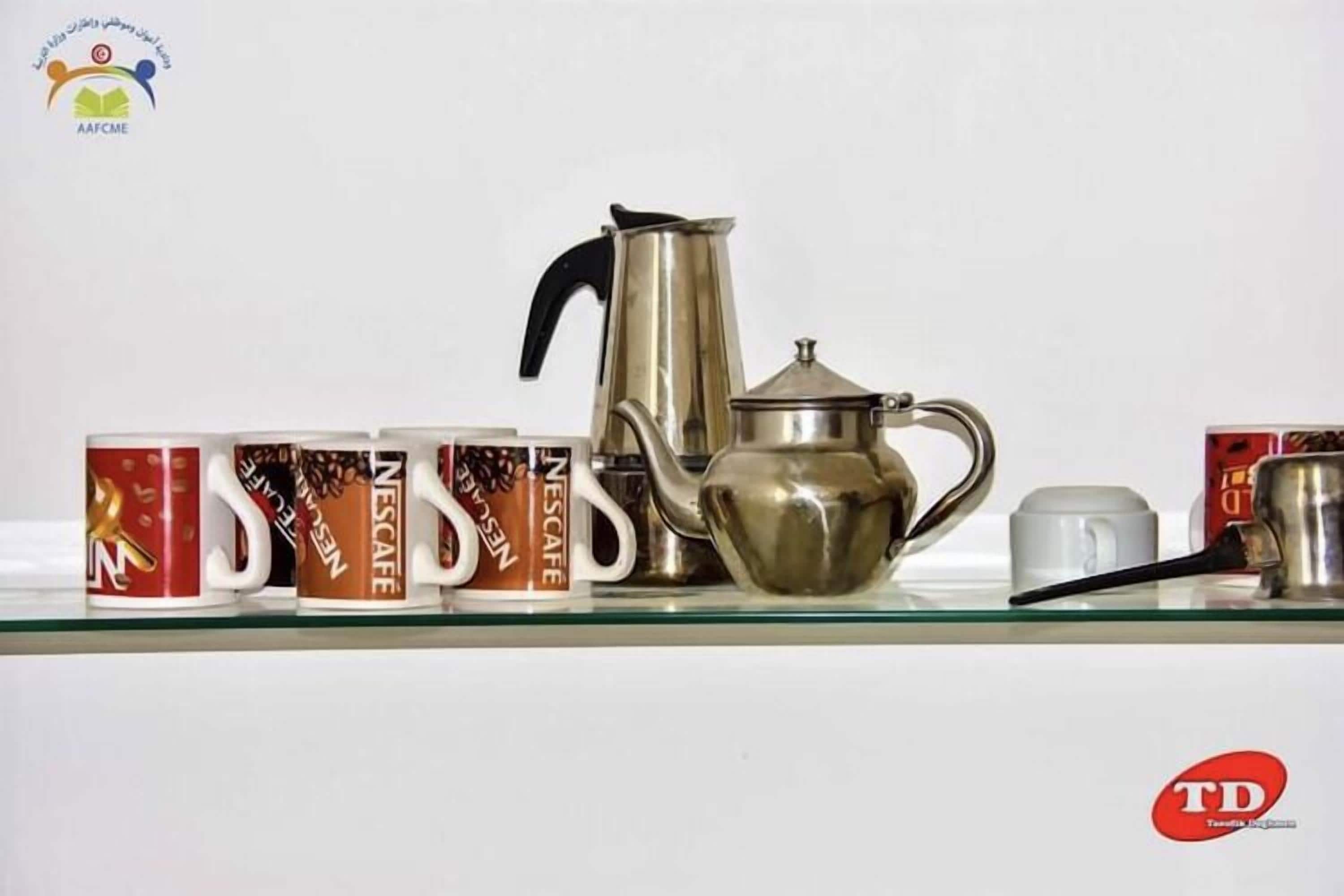 A coffee maker, a teapot, and four Nescafé cups on a glass shelf.