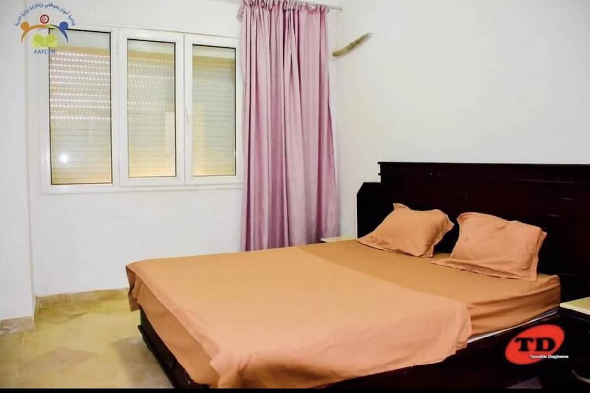 A bedroom with a bed, a window with blinds, a curtain, and a nightstand.