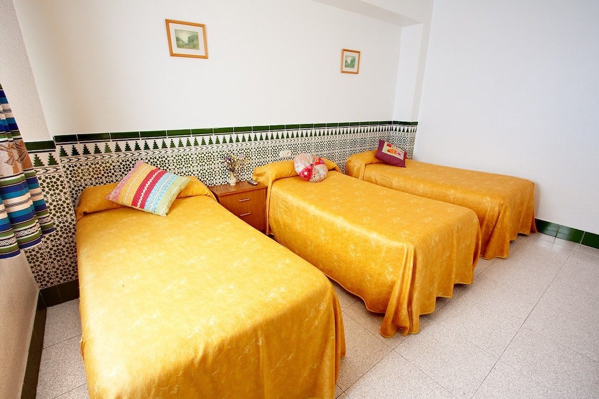 Une chambre d’hôtel avec trois lits recouverts de couvre-lits jaunes, une tête de lit à motifs et deux tableaux encadrés accrochés au mur.