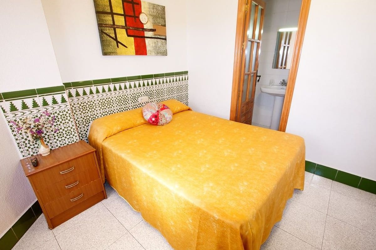 Une chambre à coucher avec un lit recouvert d’une couverture jaune, une table de chevet en bois dotée de tiroirs, un vase décoratif et un mur orné d’un tableau aux motifs géométriques.