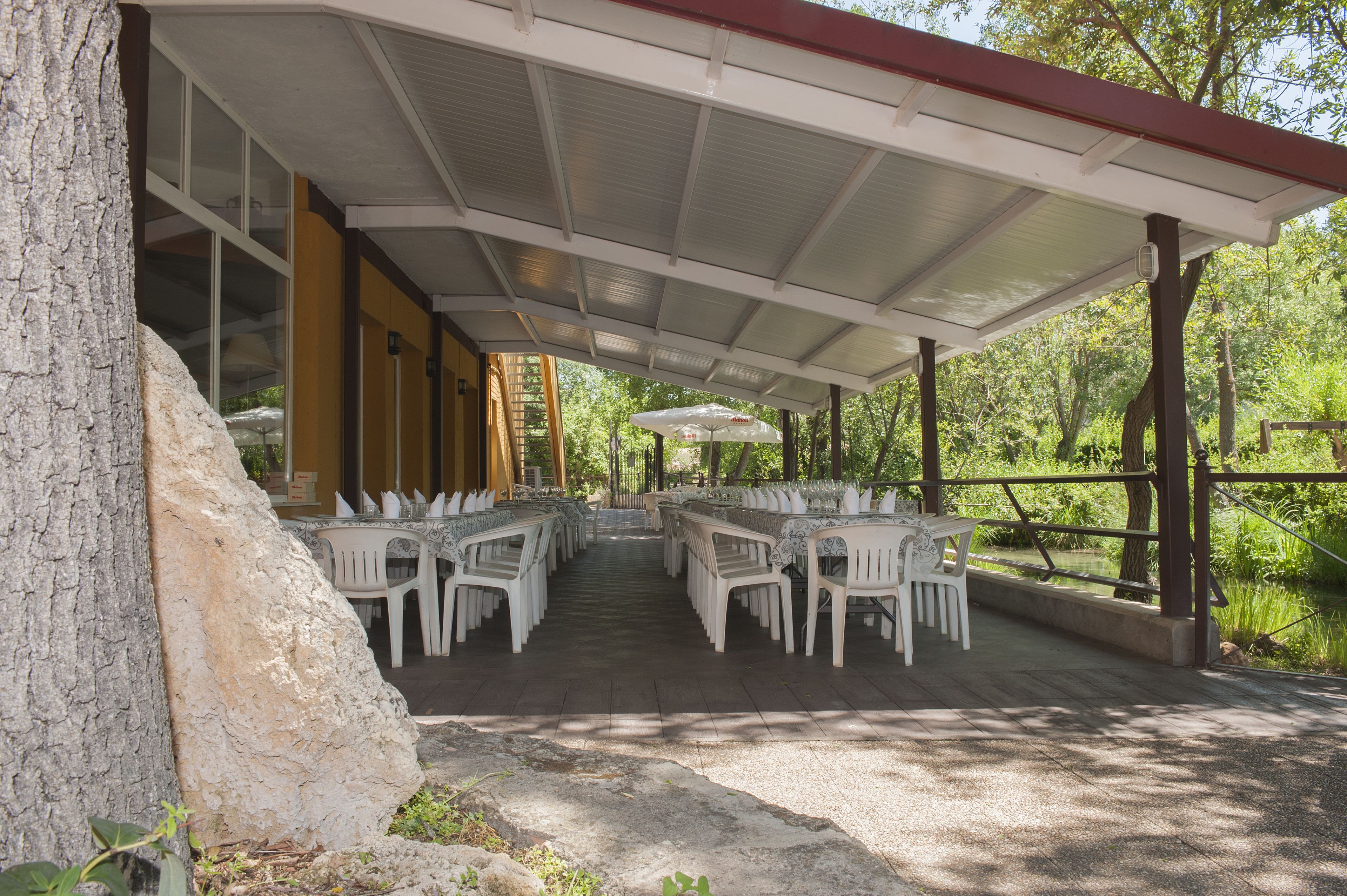 Une terrasse extérieure couverte, dotée de chaises et de tables en plastique blanc, entourée d’arbres et d’un étang.