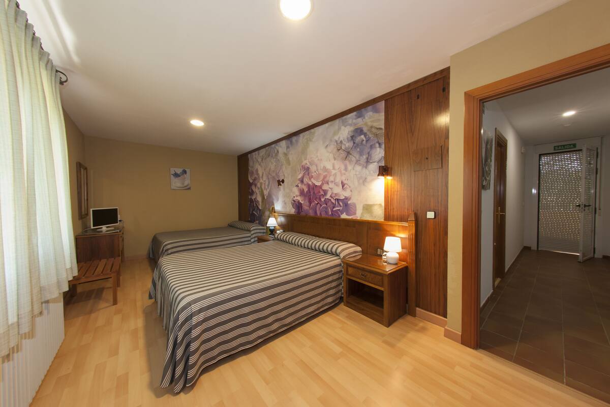 Une chambre d’hôtel avec deux lits, une table de chevet en bois avec une lampe, un tableau au mur et une vue sur le couloir.