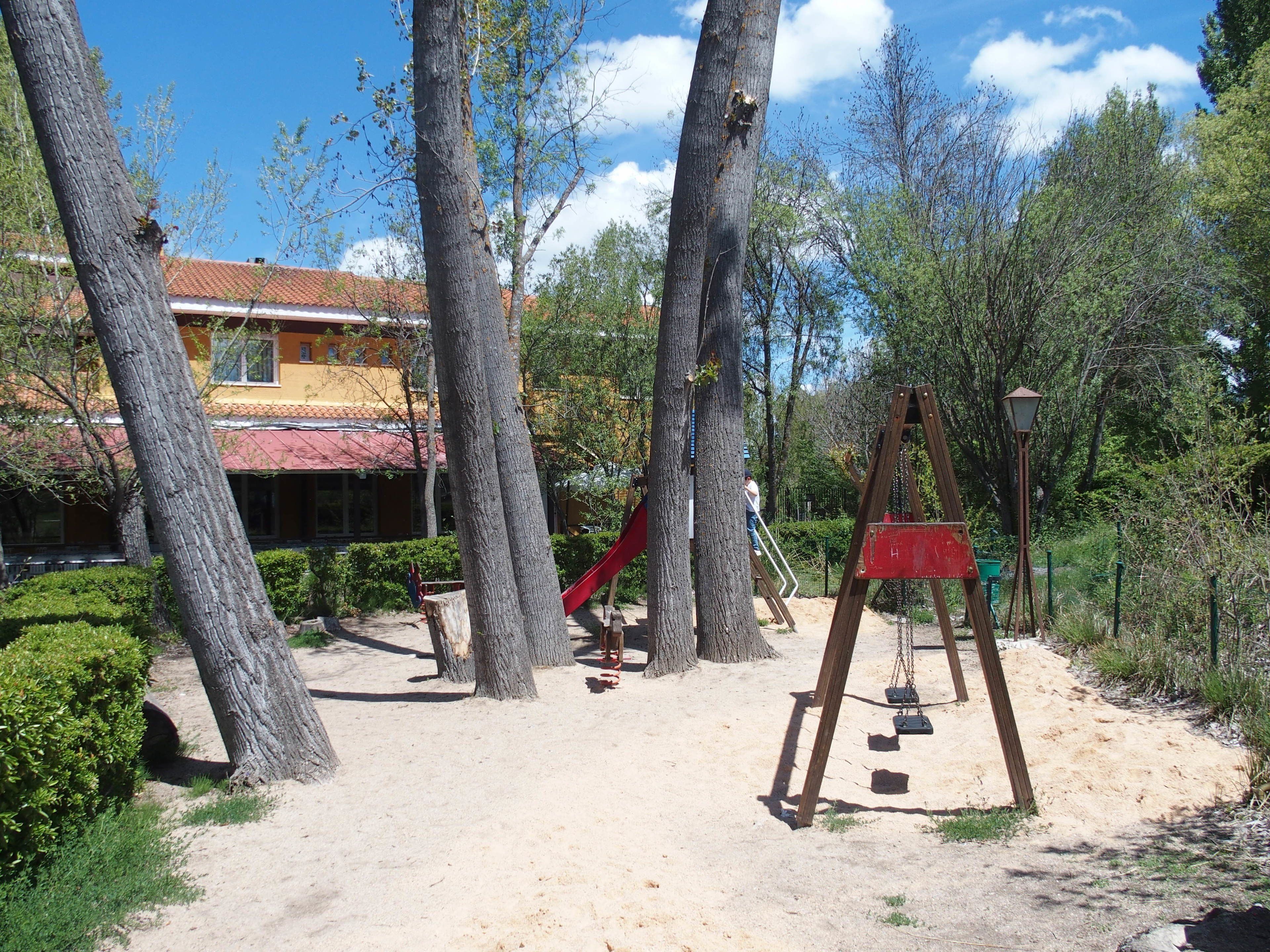 Un parque infantil con tobogán y columpios, rodeado de árboles y un edificio con techo rojo.