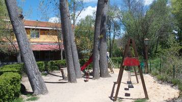 Un parque infantil con tobogán y columpios, rodeado de árboles y un edificio con techo rojo.