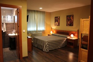 A hotel room with a bed, bedside tables, a window with curtains, a radiator, and a bathroom visible through an open door. - Hotel Restaurante El Tablazo (Villalba de la Sierra)