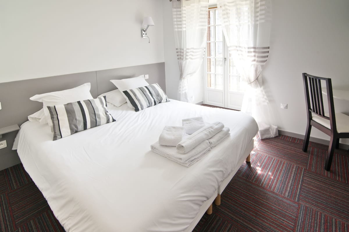 Une chambre d’hôtel avec un grand lit, du linge de lit blanc, des coussins à rayures, une chaise et une fenêtre avec des rideaux.