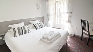 Una cama bien hecha con sábanas blancas y almohadas a rayas, una mesita con toallas dobladas y una silla.