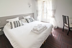 Una cama bien hecha con sábanas blancas y almohadas a rayas, una mesita con toallas dobladas y una silla.