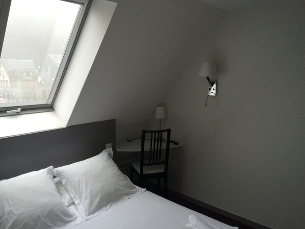 Habitación de hotel con una cama, un escritorio y una silla. Cuenta con un tragaluz sobre la cama y una lámpara de pared.