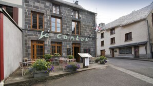 Un edificio de piedra con un cartel que dice "Le Cantou". Hay mesas y sillas al aire libre, y macetas con flores.