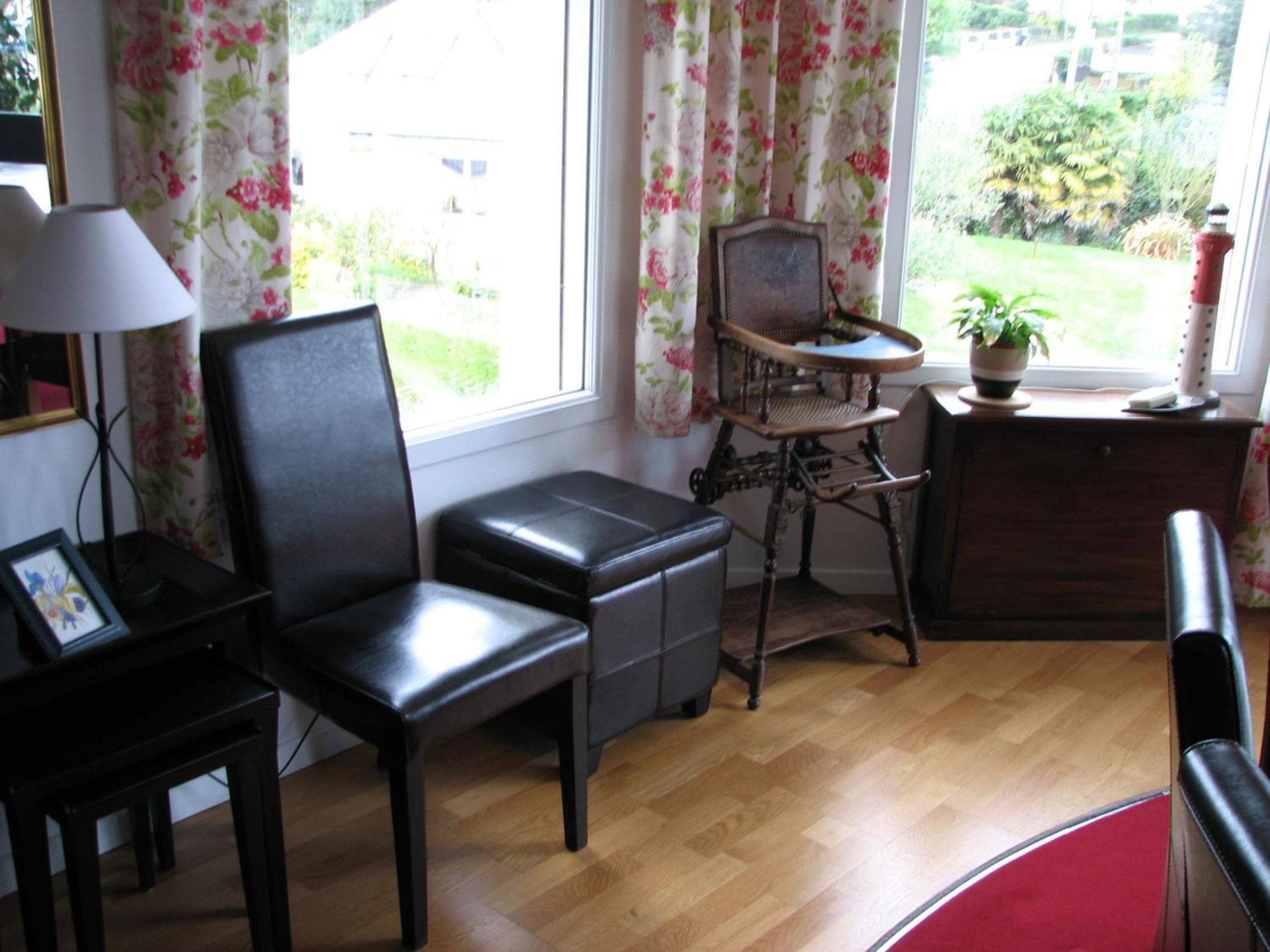 A room with a wooden floor, a black leather chair, a small wooden table, a lamp, and a window with floral curtains.