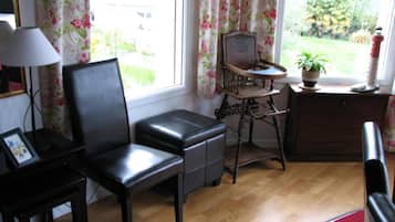 Una habitación con suelo de madera, un sillón de cuero negro, una pequeña mesa de madera, una lámpara y una ventana con cortinas florales.