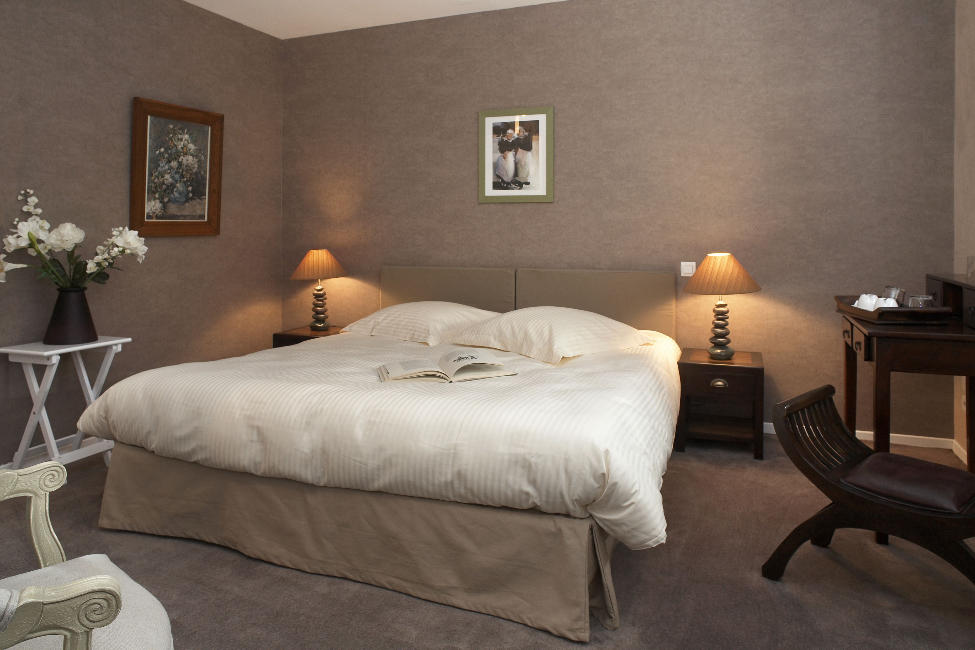 Une chambre à coucher bien rangée, avec un grand lit, deux tables de chevet avec des lampes, une chaise et une petite table sur laquelle est posé un vase de fleurs.