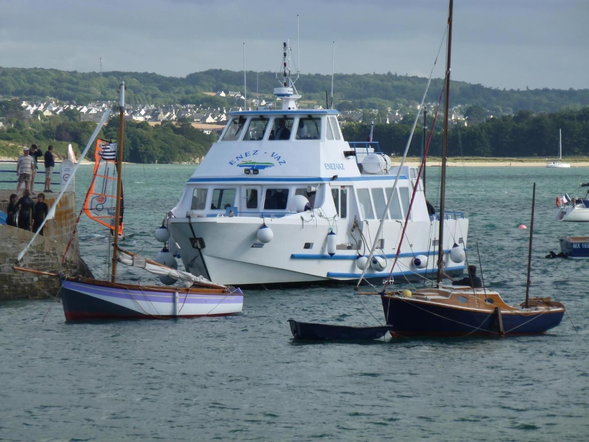 A white boat with blue trim and the name "ENEZ - VAZ" on its side, docked at a pier with other boats.