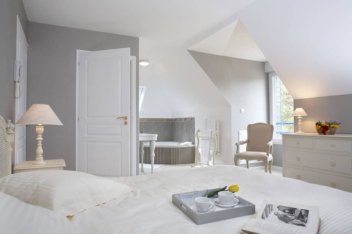 Un dormitorio con una cama grande, una mesita de noche con una lámpara, una bandeja con tazas y un libro, y vistas a un baño con bañera y sillón.