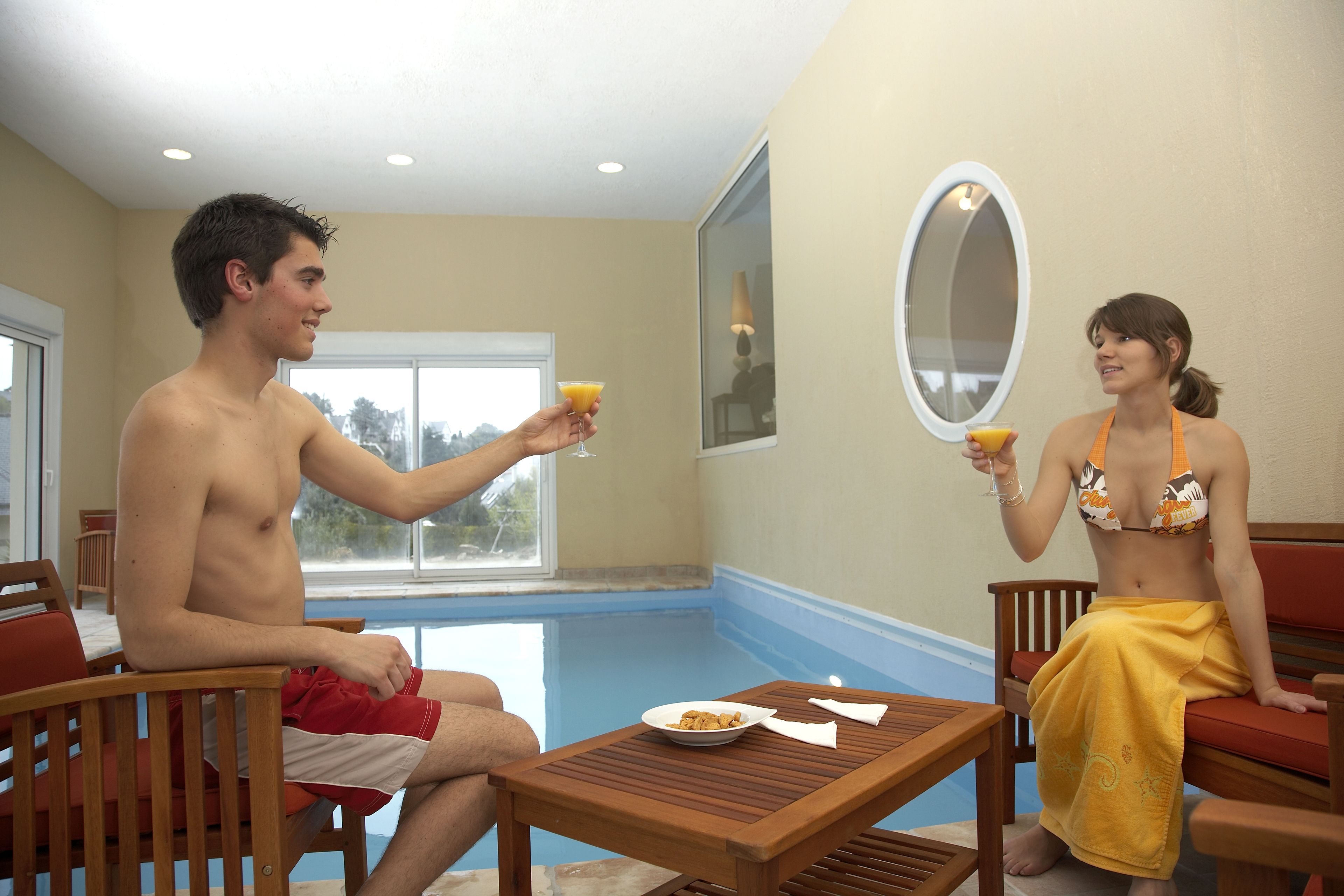 A room with a pool, a wooden table with a plate and napkin, and two people sitting on chairs.