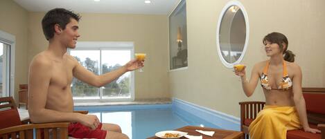 A room with a pool, a wooden table with a plate and napkin, and two people sitting on chairs.