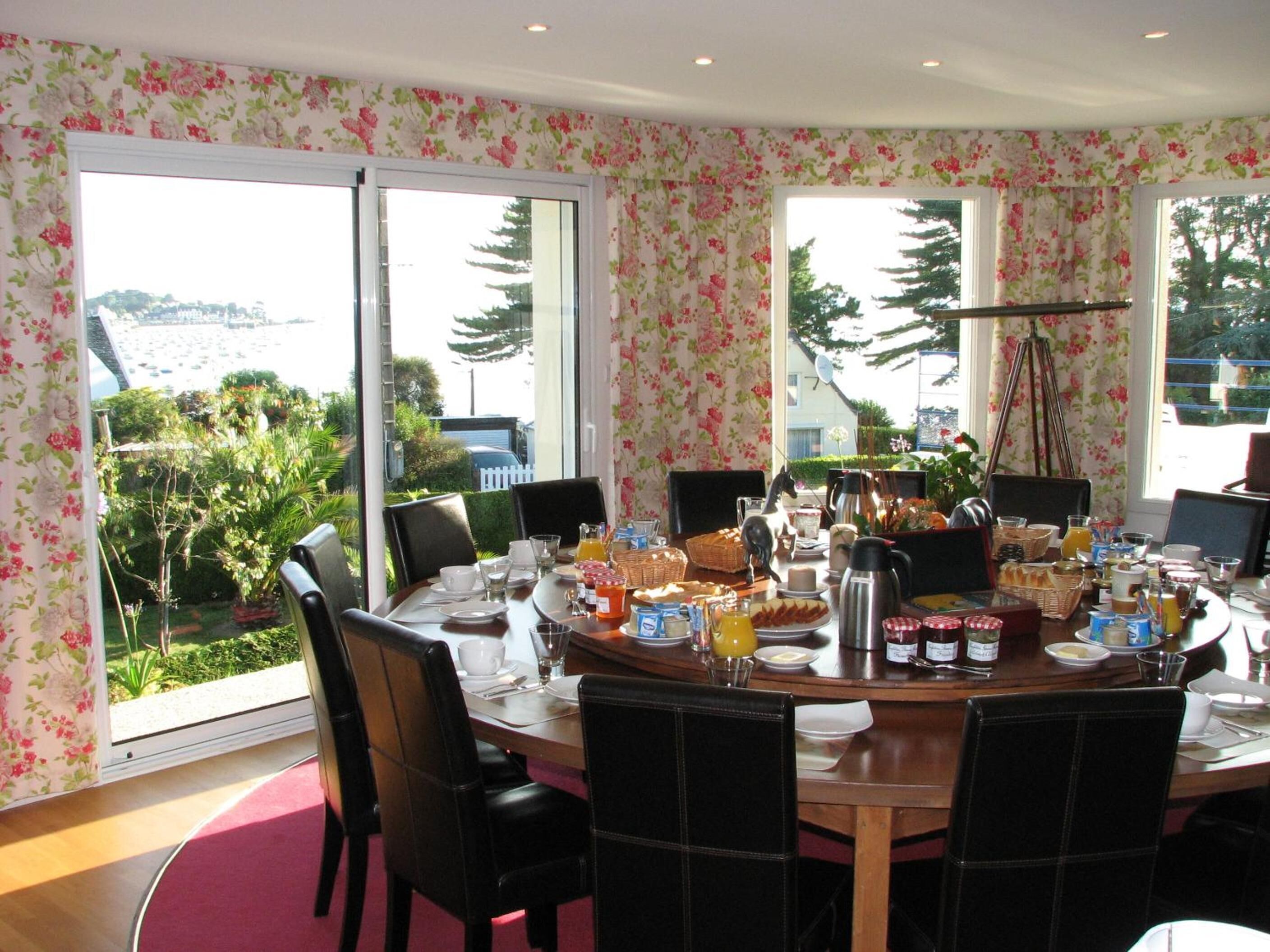 Un comedor con una mesa grande dispuesta para el desayuno, rodeada de sillas, y vistas a zonas verdes y un paisaje lejano a través de amplios ventanales.