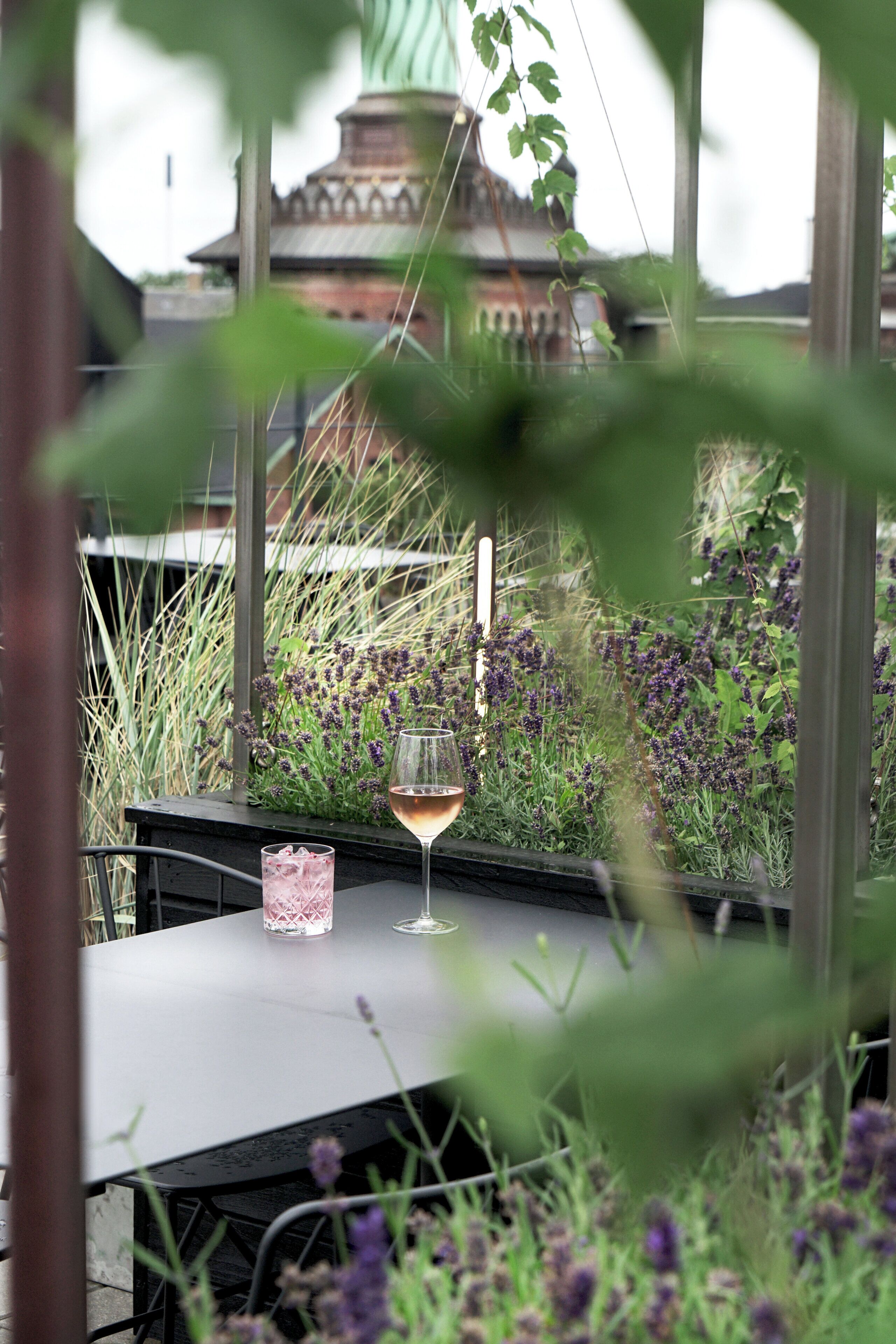 Un tavolo con un bicchiere di vino e un drink, immerso nel verde e con vista su un edificio.