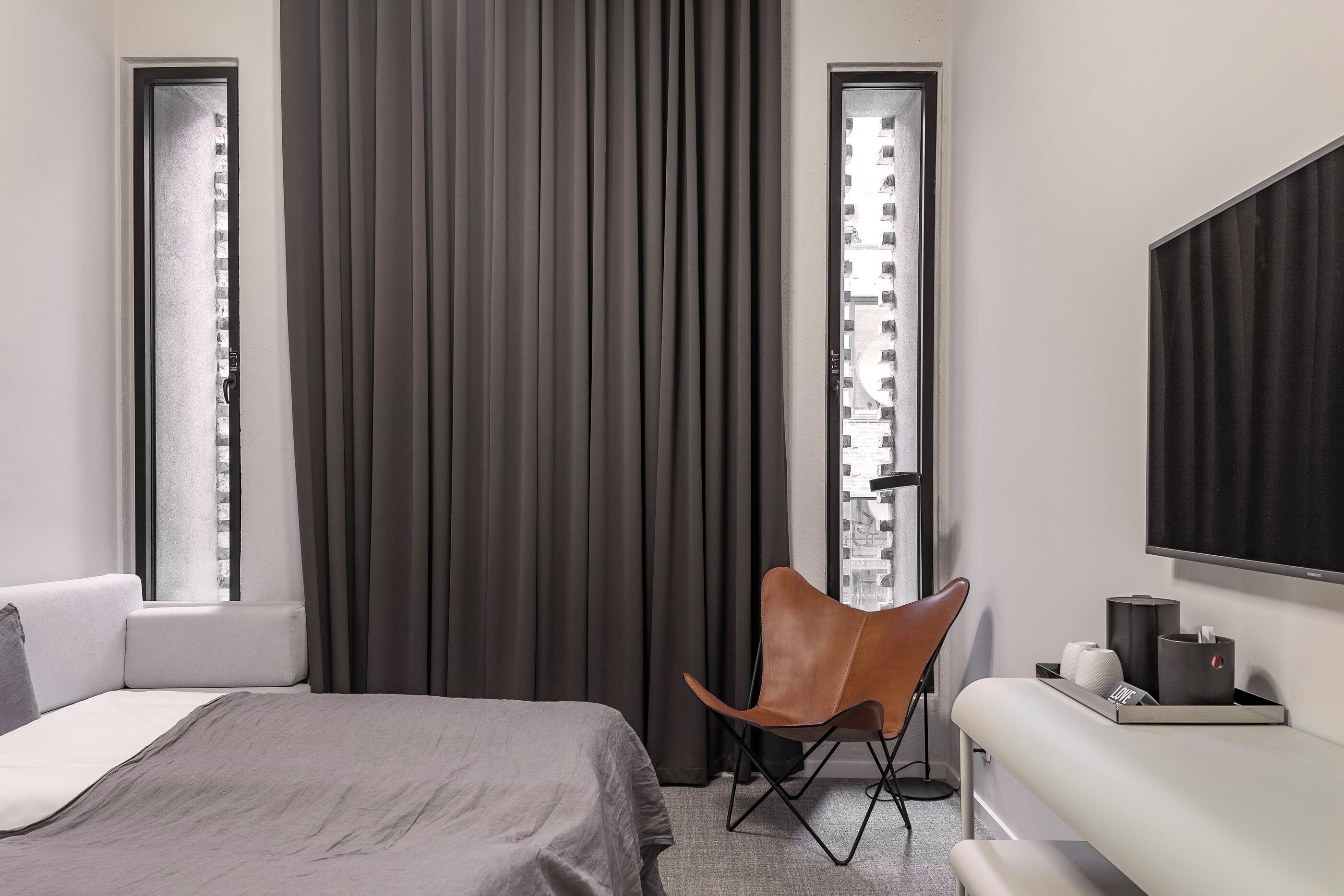 Une chambre d’hôtel moderne équipée d’un lit, d’un coin salon avec un fauteuil, d’une télévision et d’une fenêtre avec des rideaux.