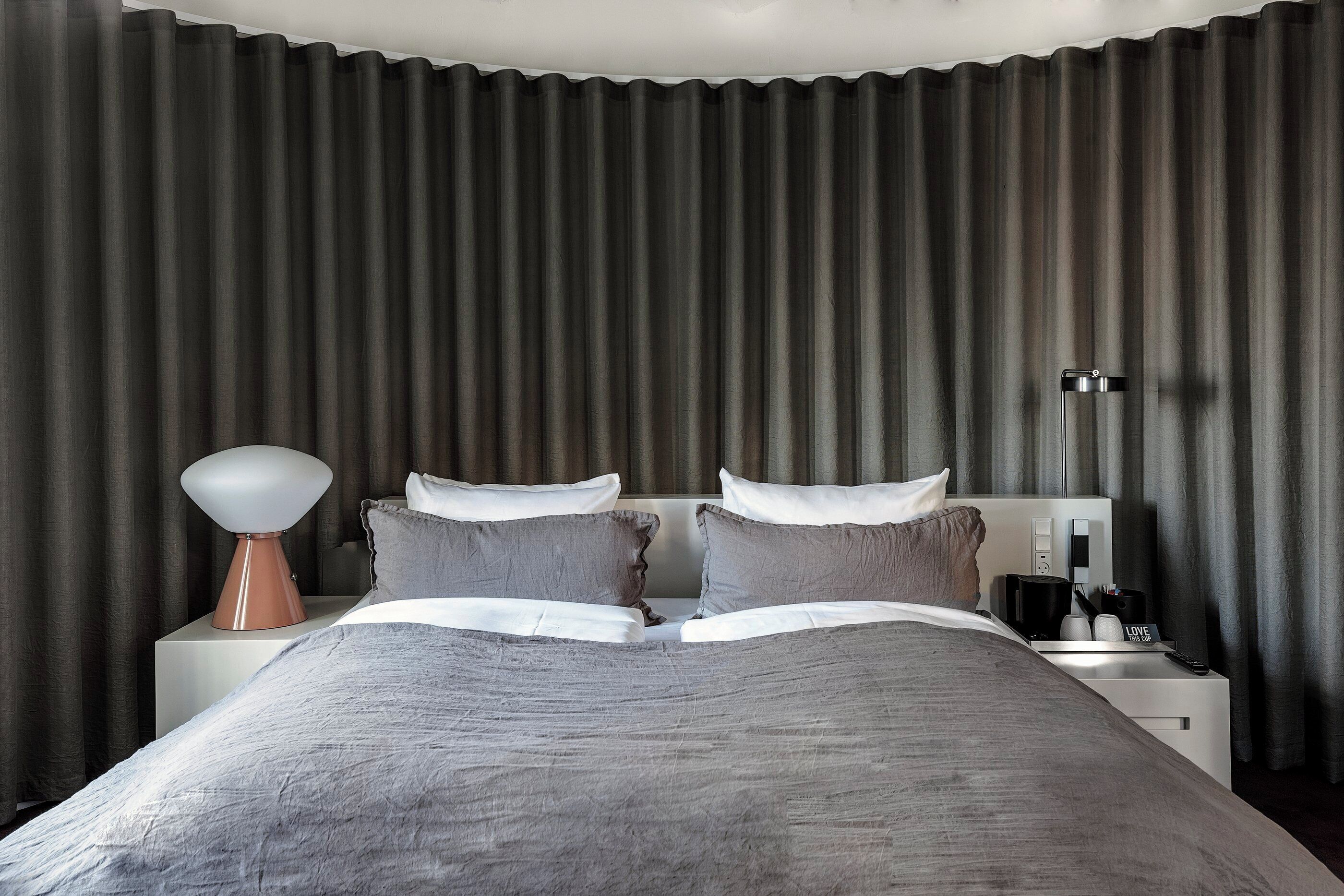 Una habitación de hotel moderna con una cama grande, una mesita de noche con lámpara y un mueble con cafetera.