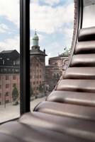 A view of a building with a green dome and a clock tower through a window with wooden blinds.